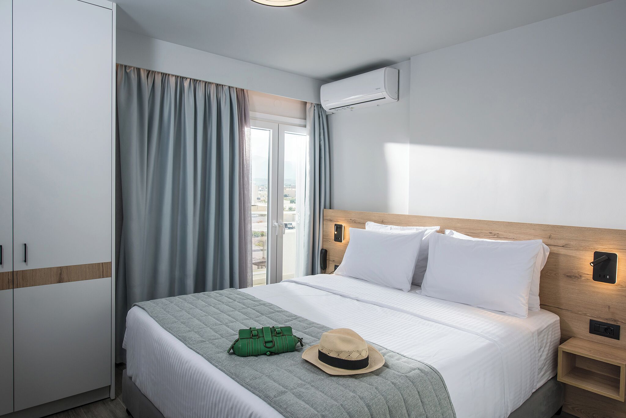 A hotel room with a bed, a wardrobe, a suitcase, a hat, and a view through the window.
