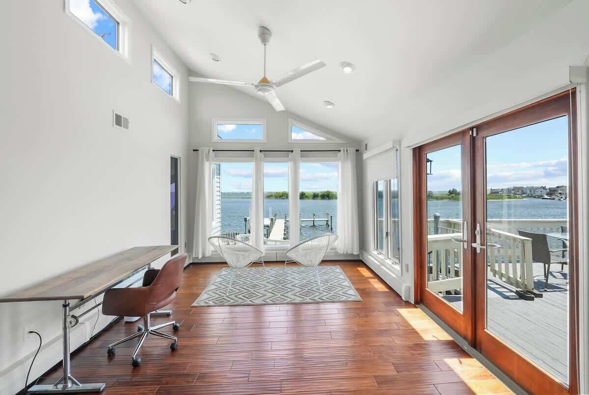 A spacious room with a wooden floor, a ceiling fan, a large window with a view of the water, and a balcony with outdoor furniture.