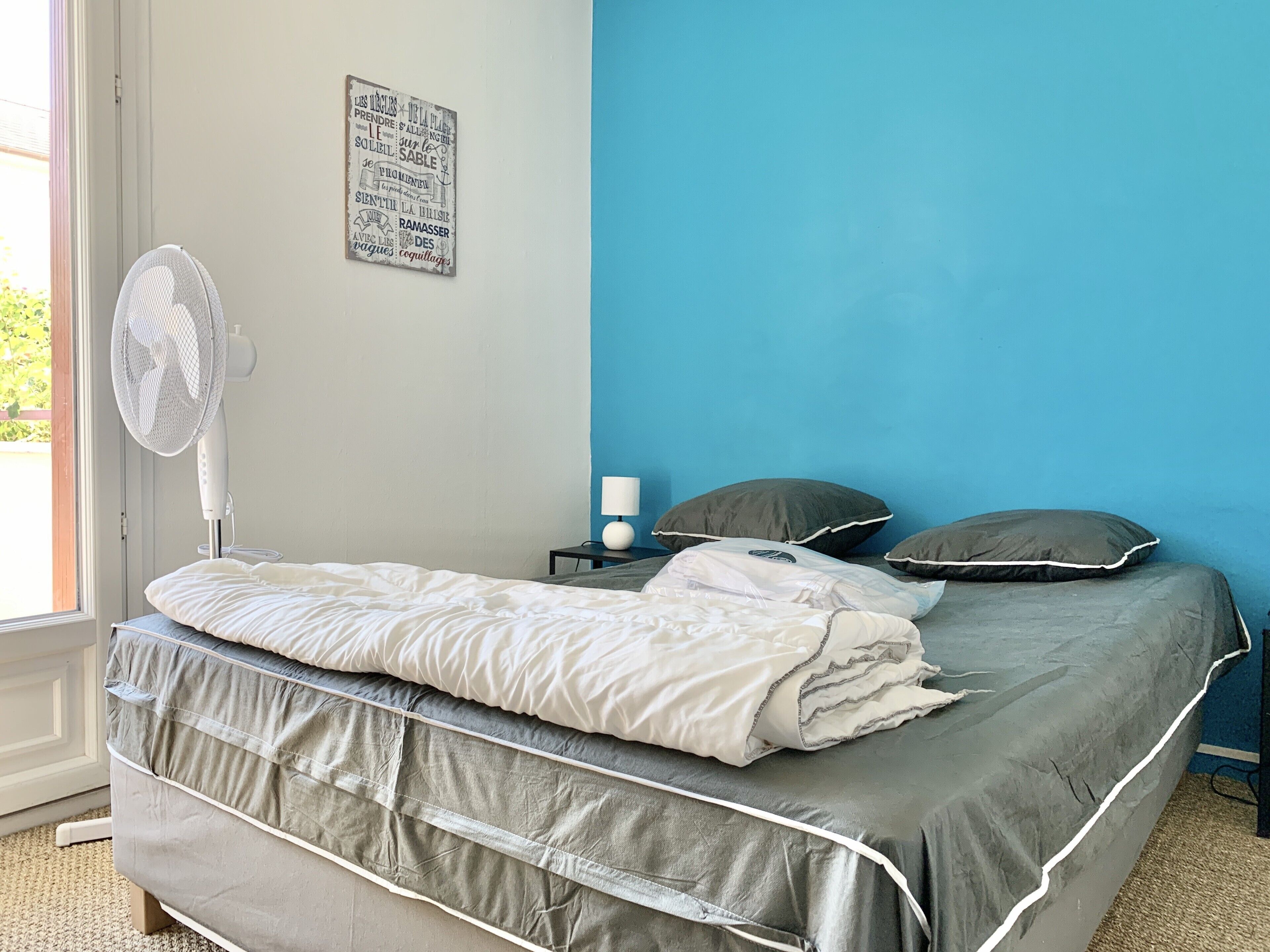 A bedroom with a bed, a nightstand, a lamp, a wall-mounted fan, and a window.