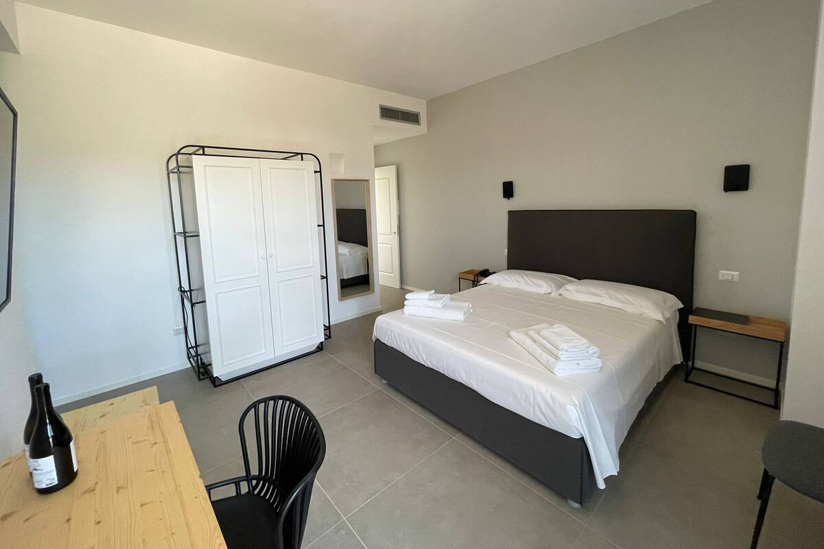 Quarto de hotel com cama, guarda-roupa, escrivaninha e uma cadeira, além de uma garrafa de vinho.