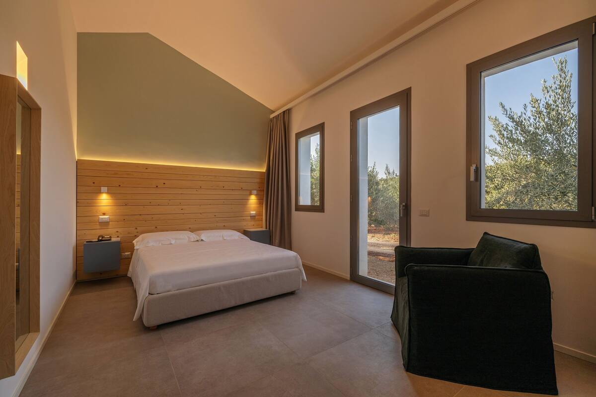 Quarto com cama, mesas de cabeceira, janela e vista para árvores.