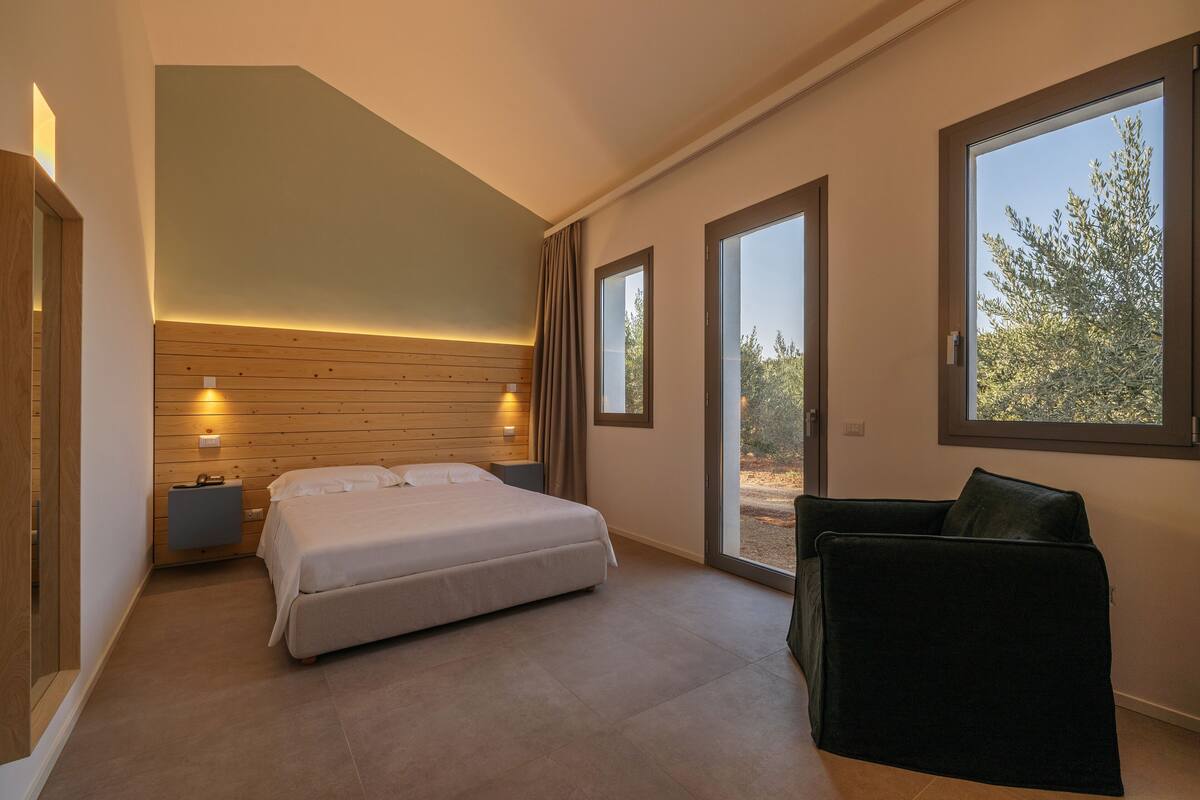 Quarto moderno com cama, mesas de cabeceira, uma cadeira e grandes janelas com vista para o exterior.