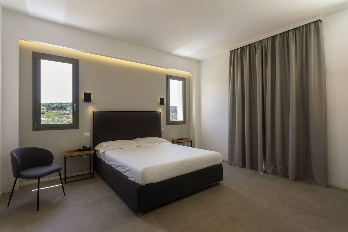 Quarto moderno com uma cama grande, duas mesas de cabeceira, uma cadeira e vista para o exterior por duas janelas.