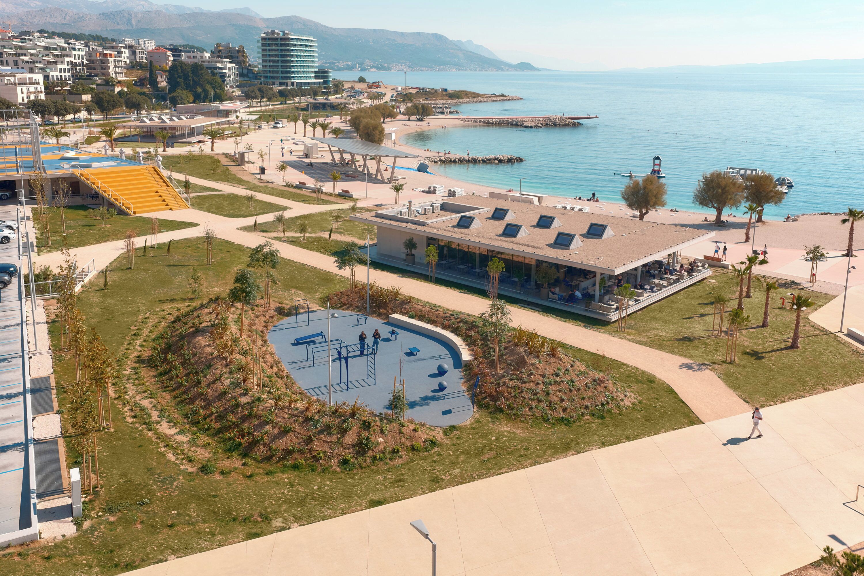 Vista aèria d'una zona costanera amb una piscina, un edifici amb teulada inclinada i un passeig marítim al costat de l'aigua.