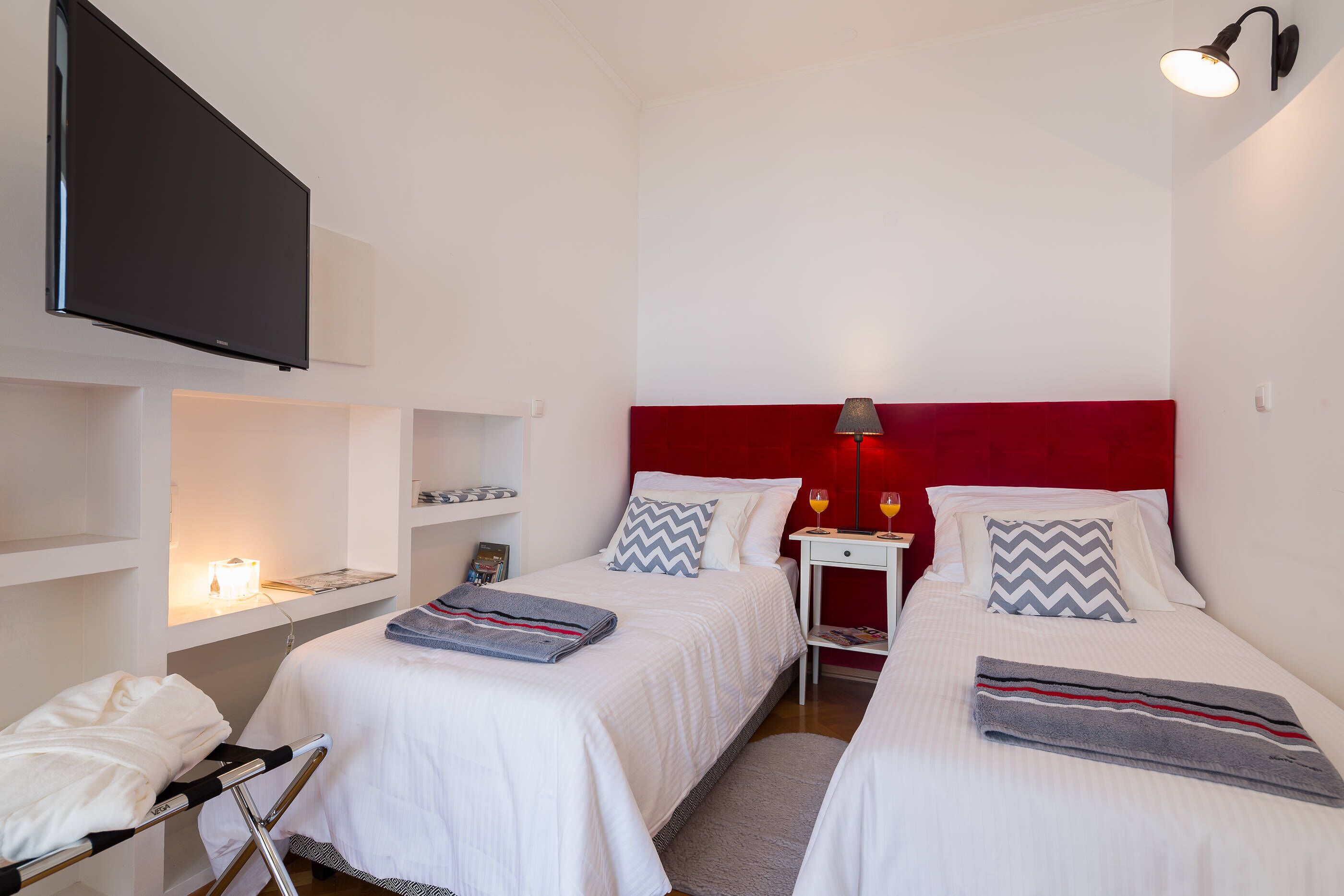 A hotel room with two beds, a TV, a bedside table with a lamp, and a shelf with a lamp.