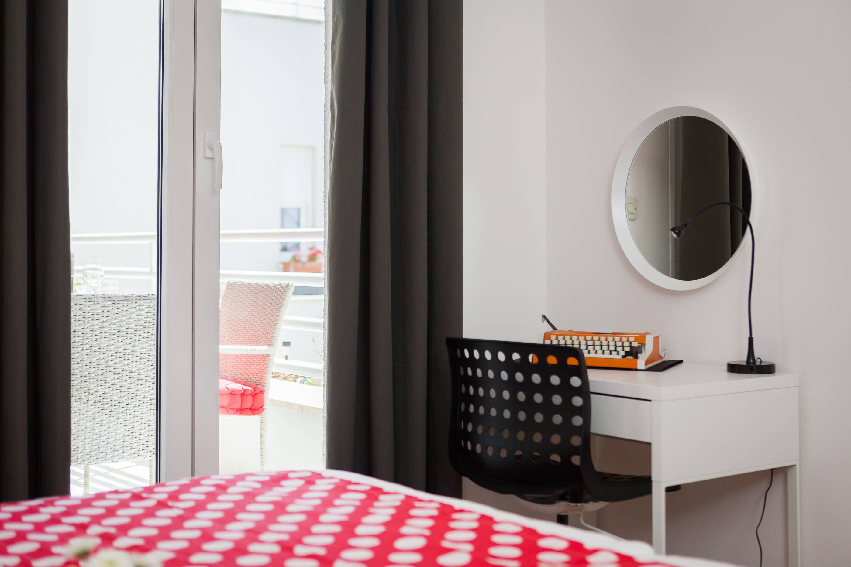 Ein Hotelzimmer mit einem Bett, einem Schreibtisch mit Stuhl, einem runden Spiegel und einem Fenster mit Vorhängen.