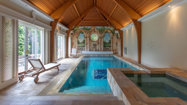 An indoor swimming pool with wooden beams, a mural, and a view through large windows. - Luxury country retreat with extensive leisure facilities (Lane End)