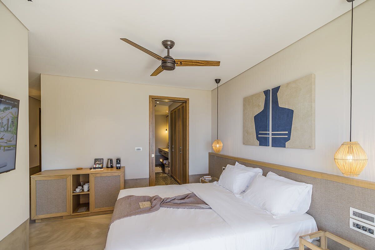 A hotel room with a bed, a ceiling fan, a bedside table, a lamp, and a painting on the wall.