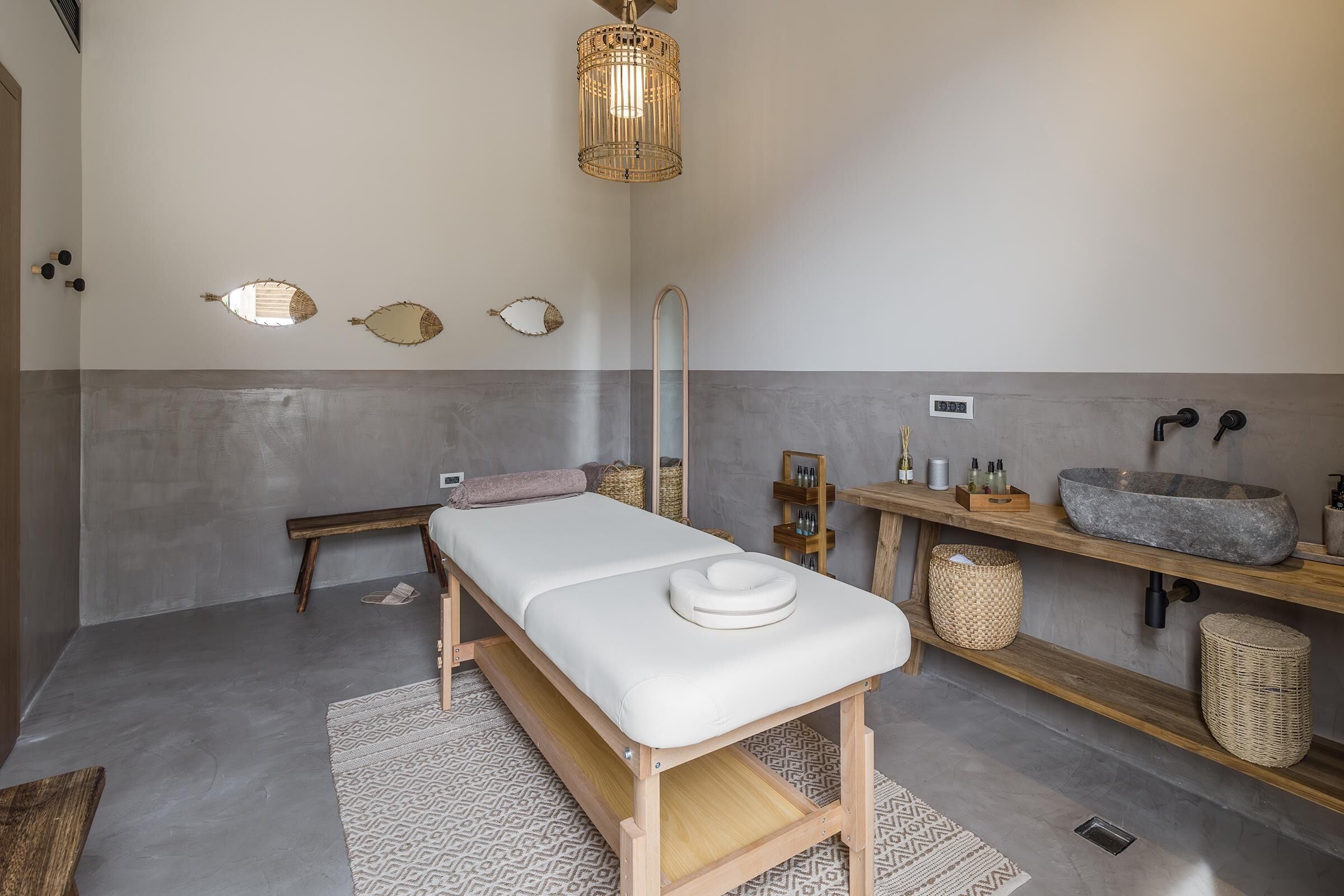 A modern spa room with a wooden massage table, a large mirror, a stone sink, and a ceiling light fixture.