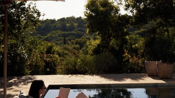A person relaxing by a poolside lounge chair with a book, a glass of water, and a basket.
