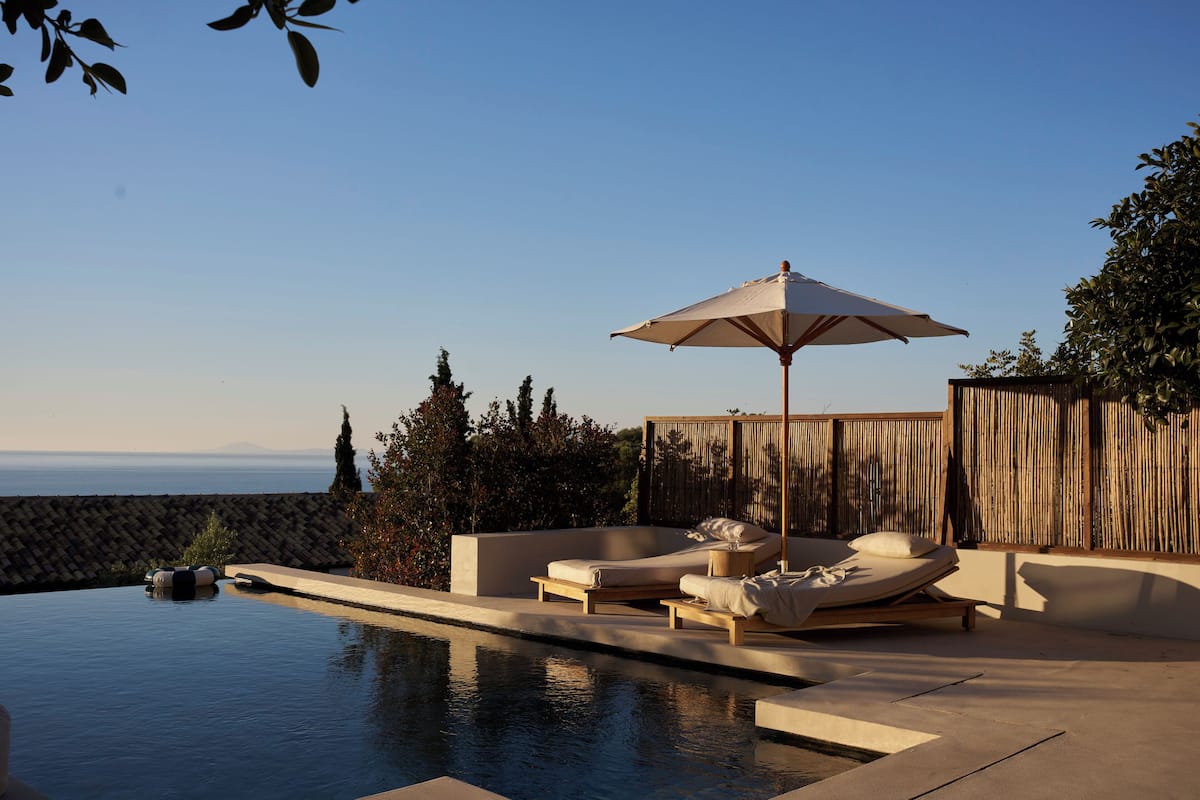 Un área de piscina con sillas de descanso y una sombrilla, rodeada por una valla de madera y vegetación.