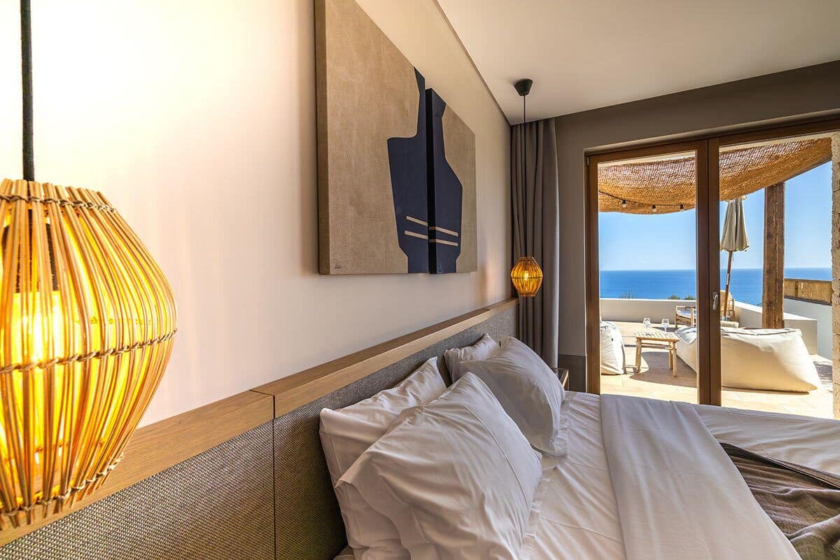 Habitación de hotel con cama, un ventanal con vista al mar, una lámpara colgante y un cuadro en la pared.