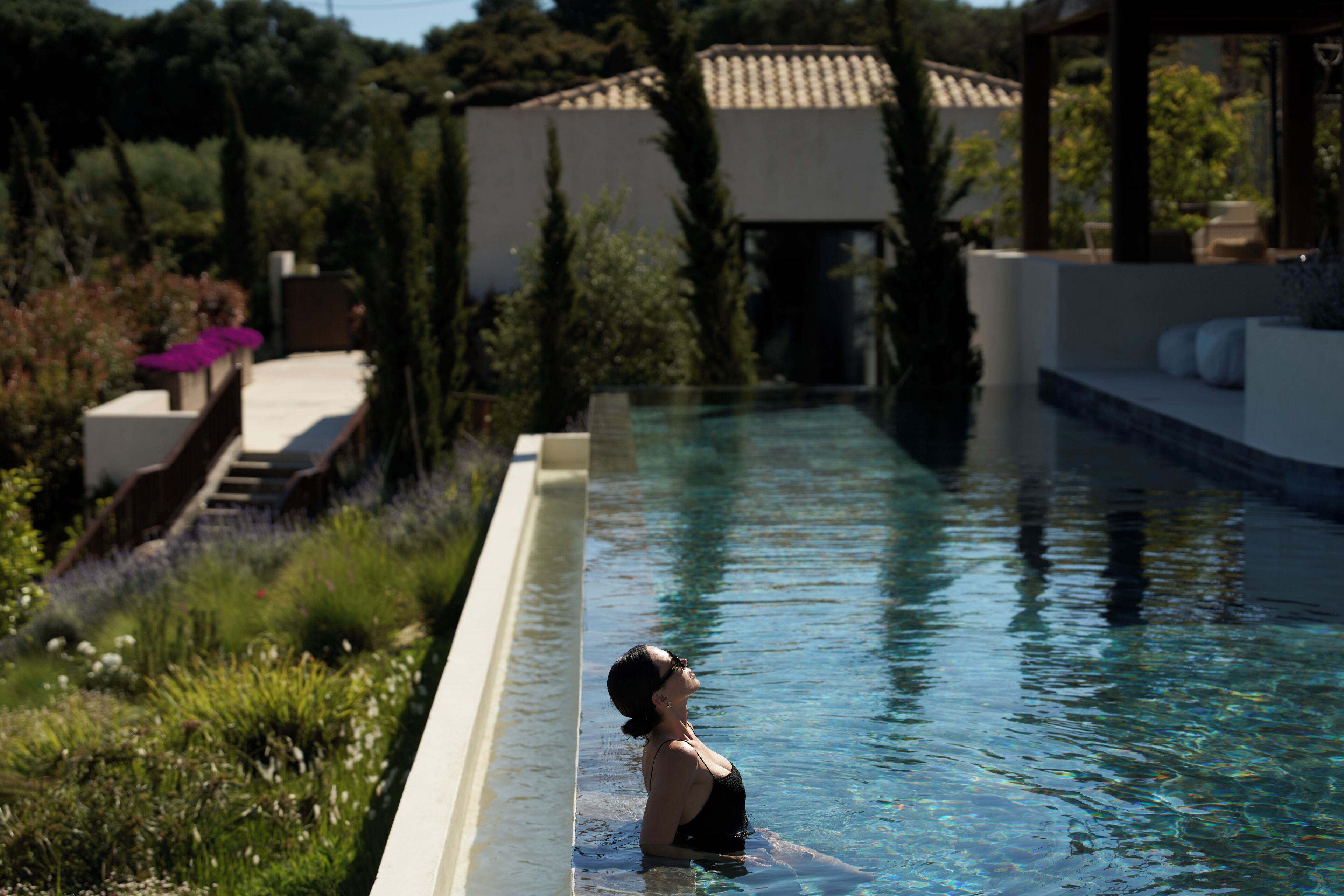 A swimming pool with a person in the water, surrounded by greenery and a building with a tiled roof.