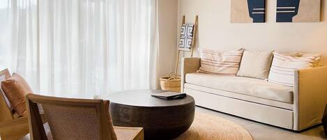 A modern living room with a round coffee table, a sofa, and a wooden chair. There are abstract paintings on the wall and a window with sheer curtains.