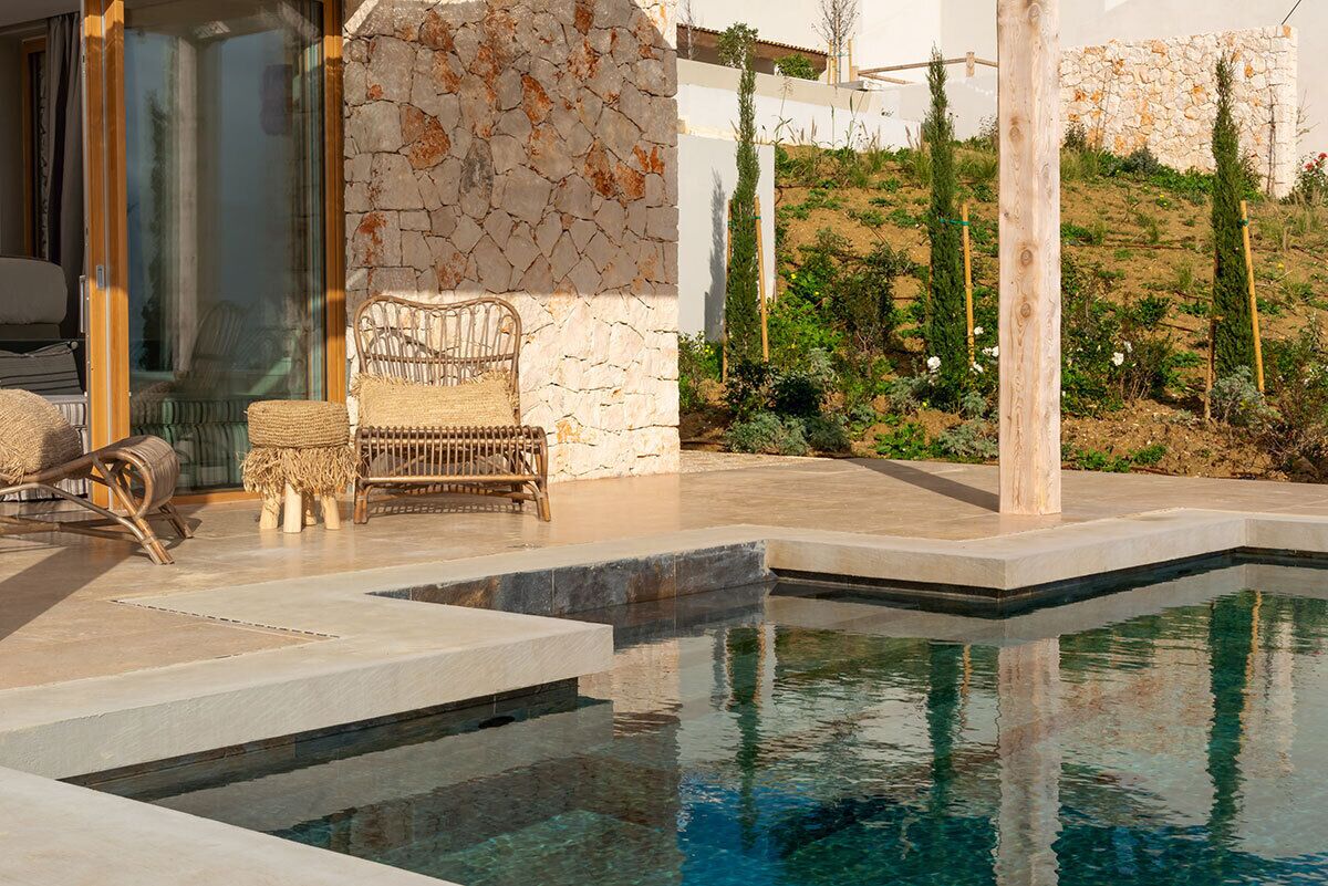 Área de piscina com paredes de pedra, uma cadeira de vime e vista para um jardim em encosta.