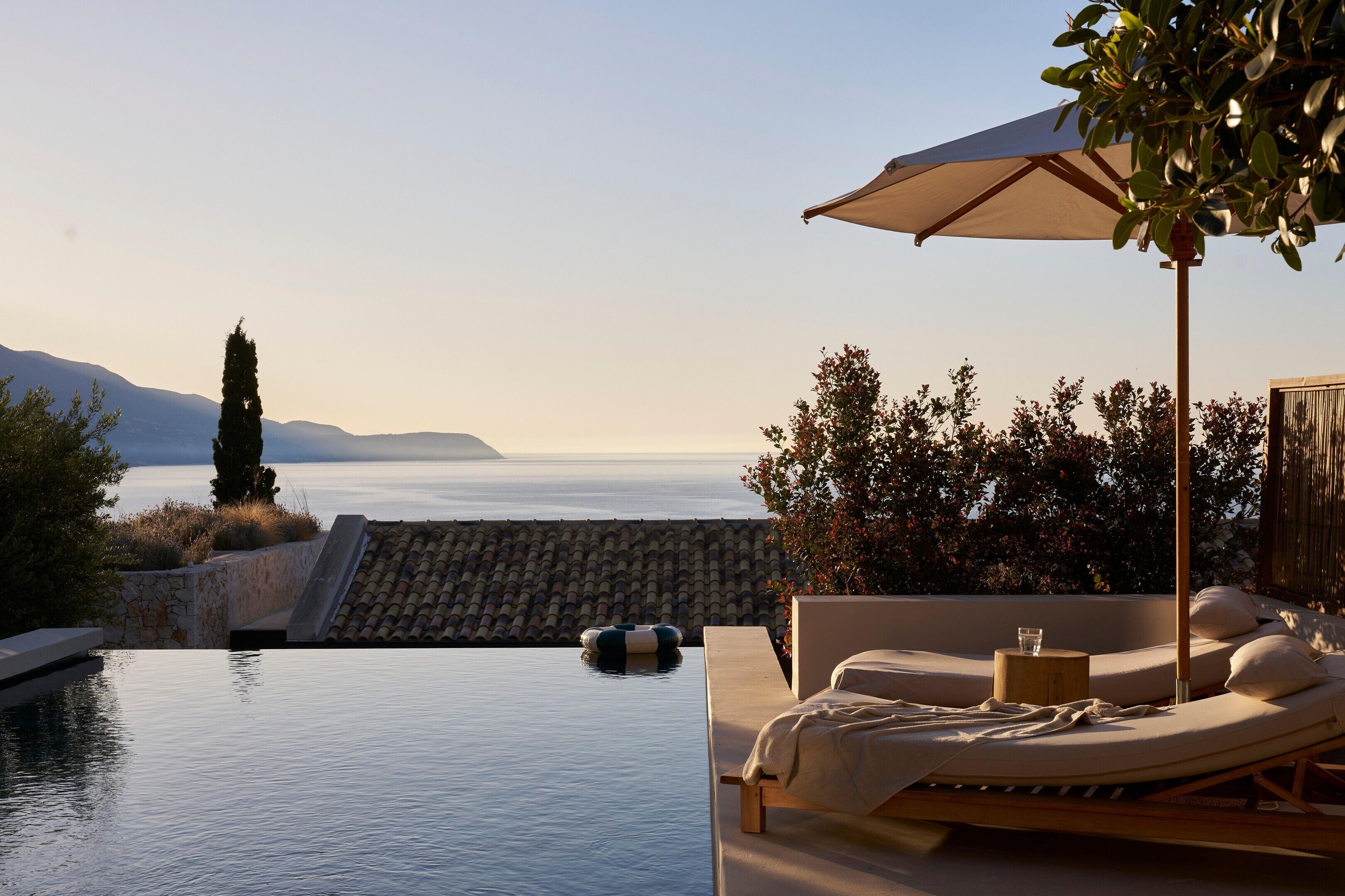 A serene outdoor lounge area with a view of the sea and mountains, a swimming pool, and a sunbed under a canopy.