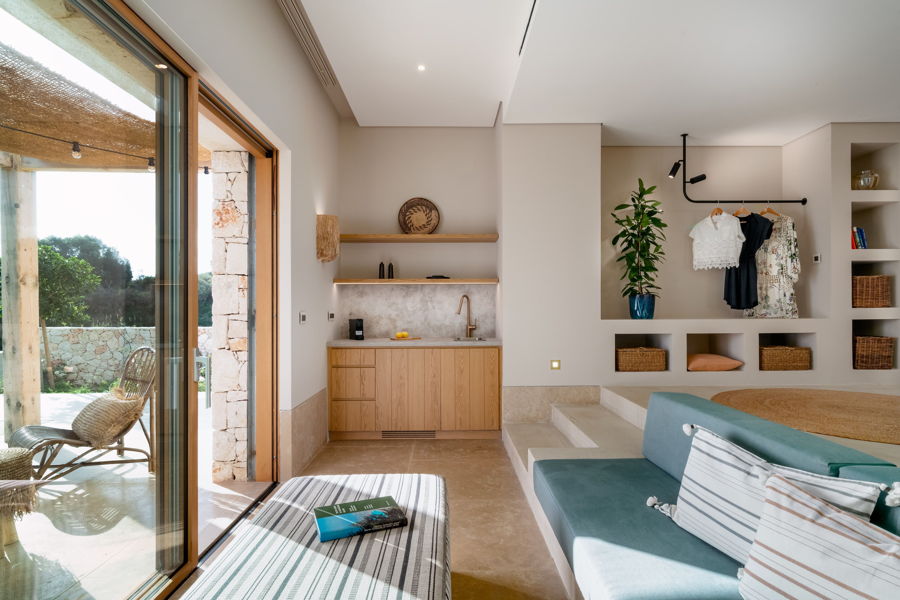 A modern interior with a glass door leading to an outdoor area, a kitchenette with wooden cabinets, a sofa with cushions, and a wall-mounted rack with clothes and baskets.