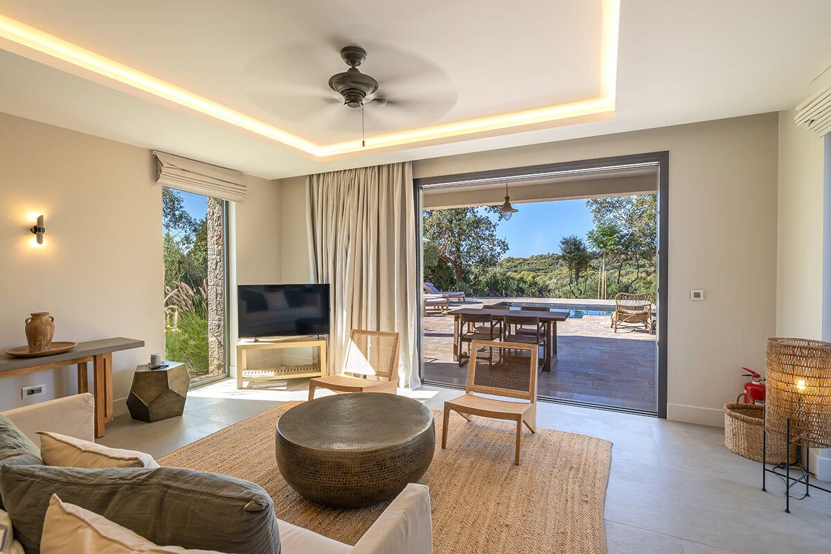 A modern living room with a flat-screen TV, comfortable seating, and a large window offering a view of an outdoor pool area.