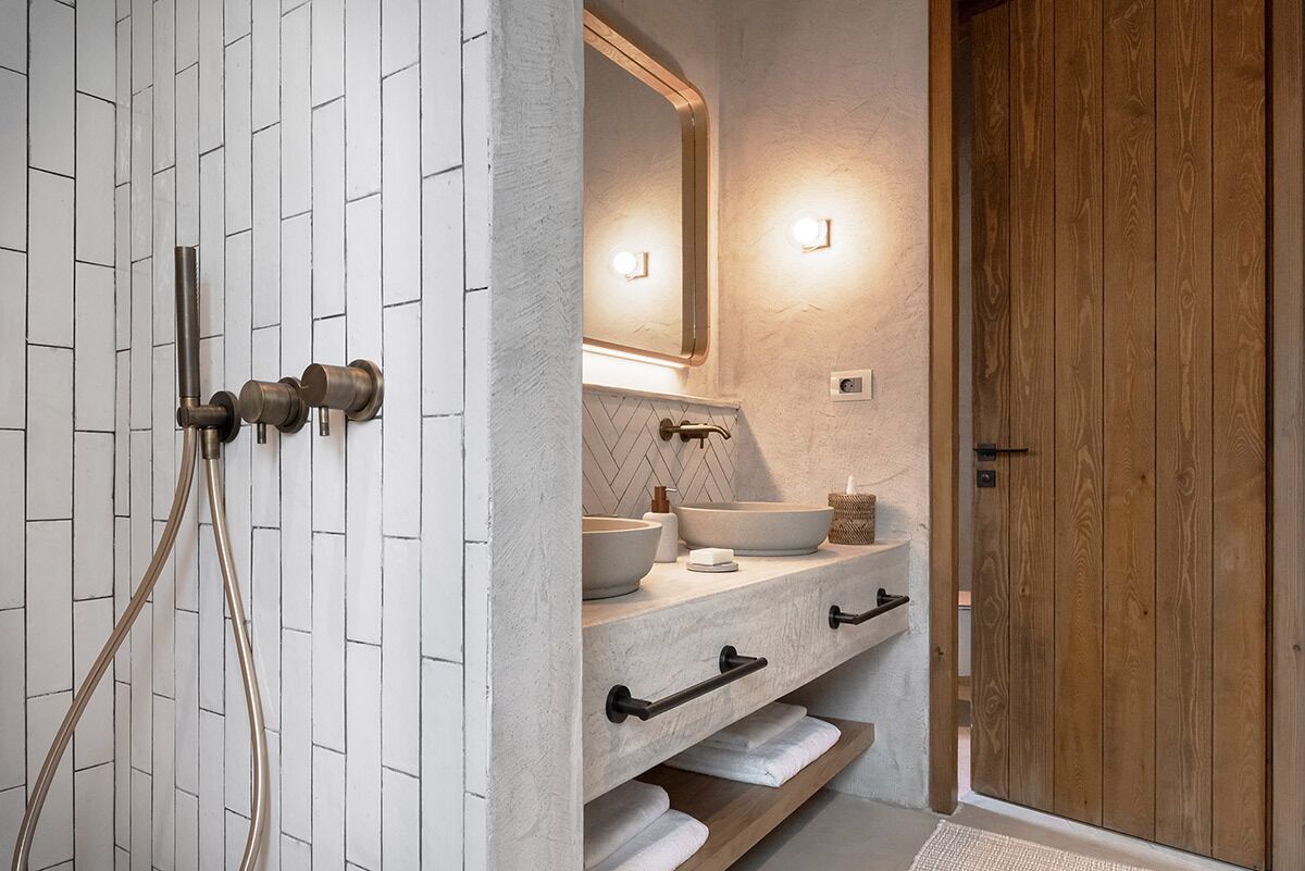 A bathroom with a white tiled wall, a wooden door, and a vanity with a mirror and two sinks.