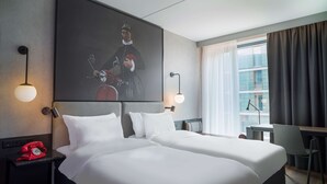 A hotel room with a large bed, a desk, a chair, a red telephone, and a painting on the wall.