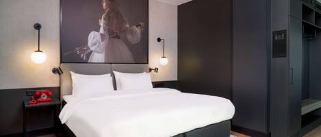 Una habitación de hotel moderna con una cama grande, una mesita de noche pequeña con un teléfono rojo, dos lámparas fijadas a la pared, un cuadro grande enmarcado y un armario empotrado.