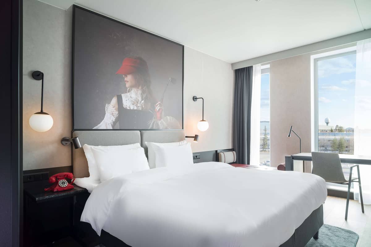 A hotel room with a large bed, a red telephone, two wall-mounted lamps, a large window with a view, and a desk with a chair.