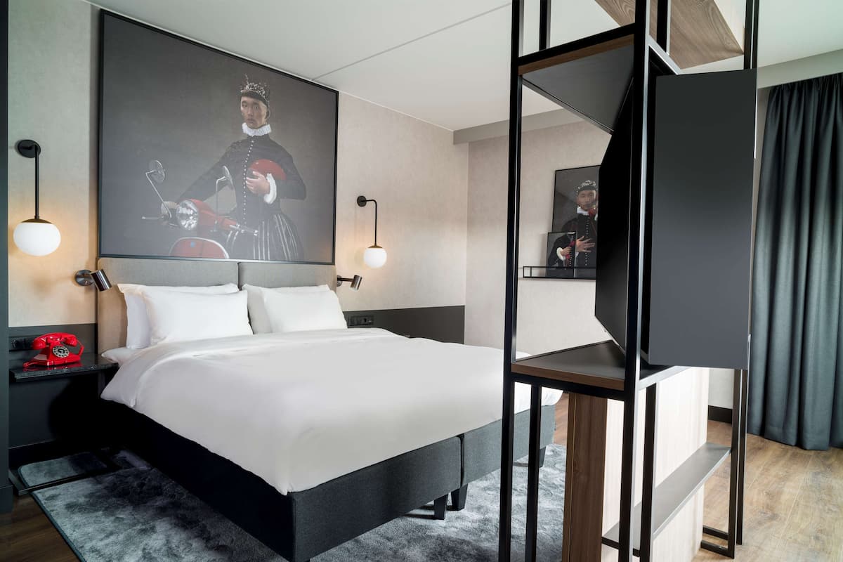 A modern hotel room with a large bed, a flat-screen TV, a red rotary phone, and a framed portrait on the wall.