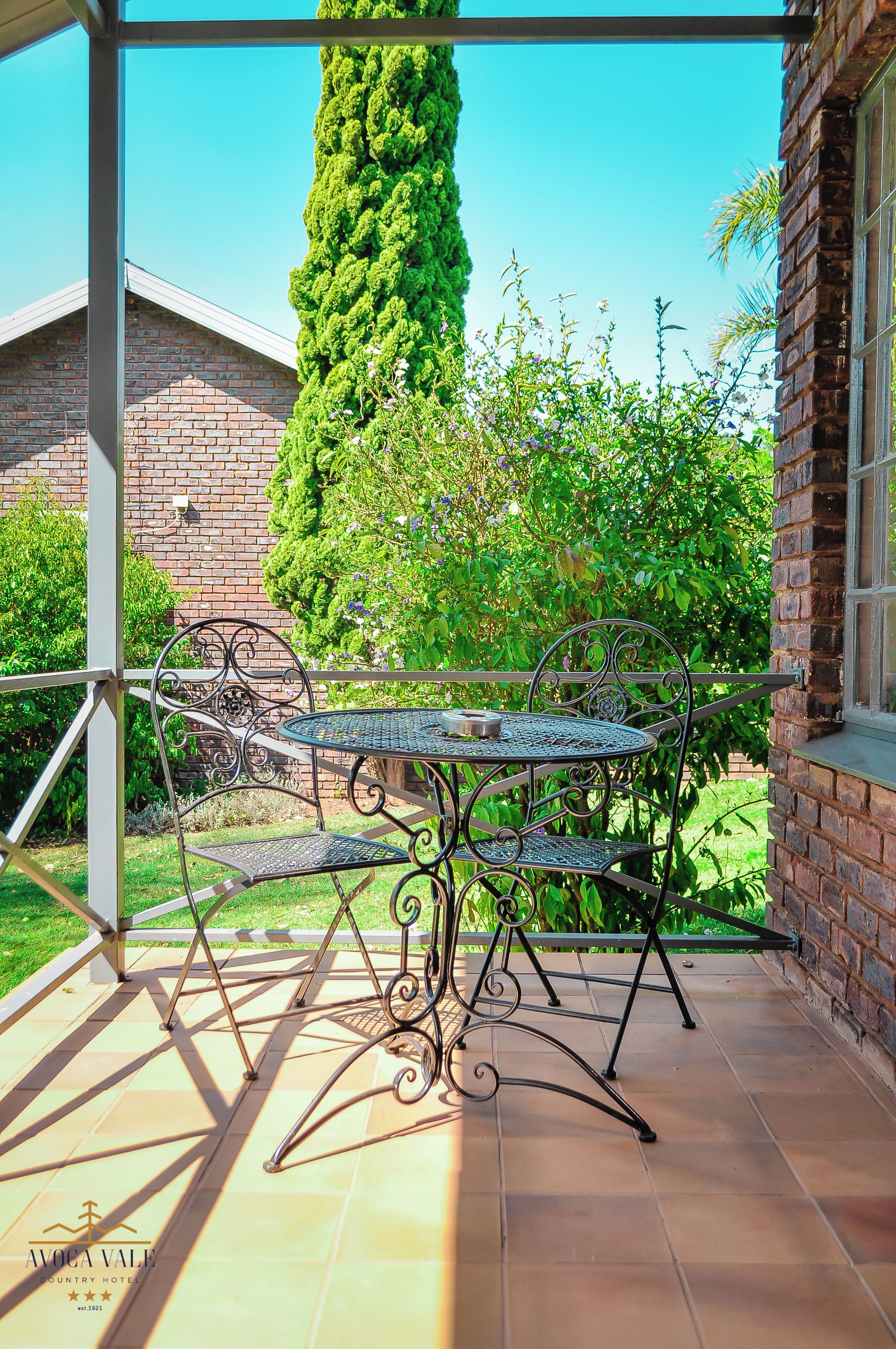 Un patio con una mesa y sillas de metal, rodeado por un muro de ladrillo y follaje.