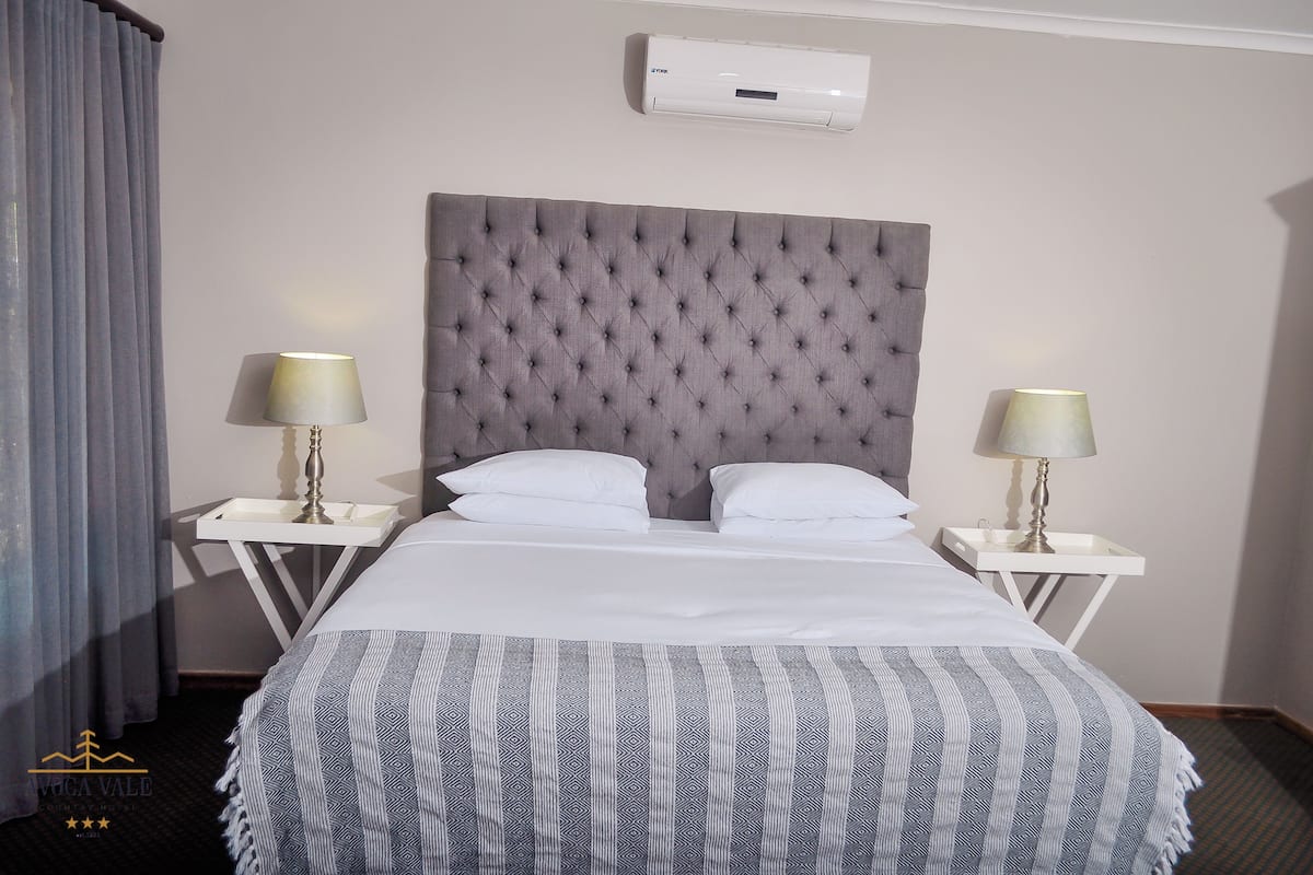 Quarto de hotel com cabeceira estofada, duas mesas de cabeceira com luminárias, cama com roupa de cama branca e manta decorativa.