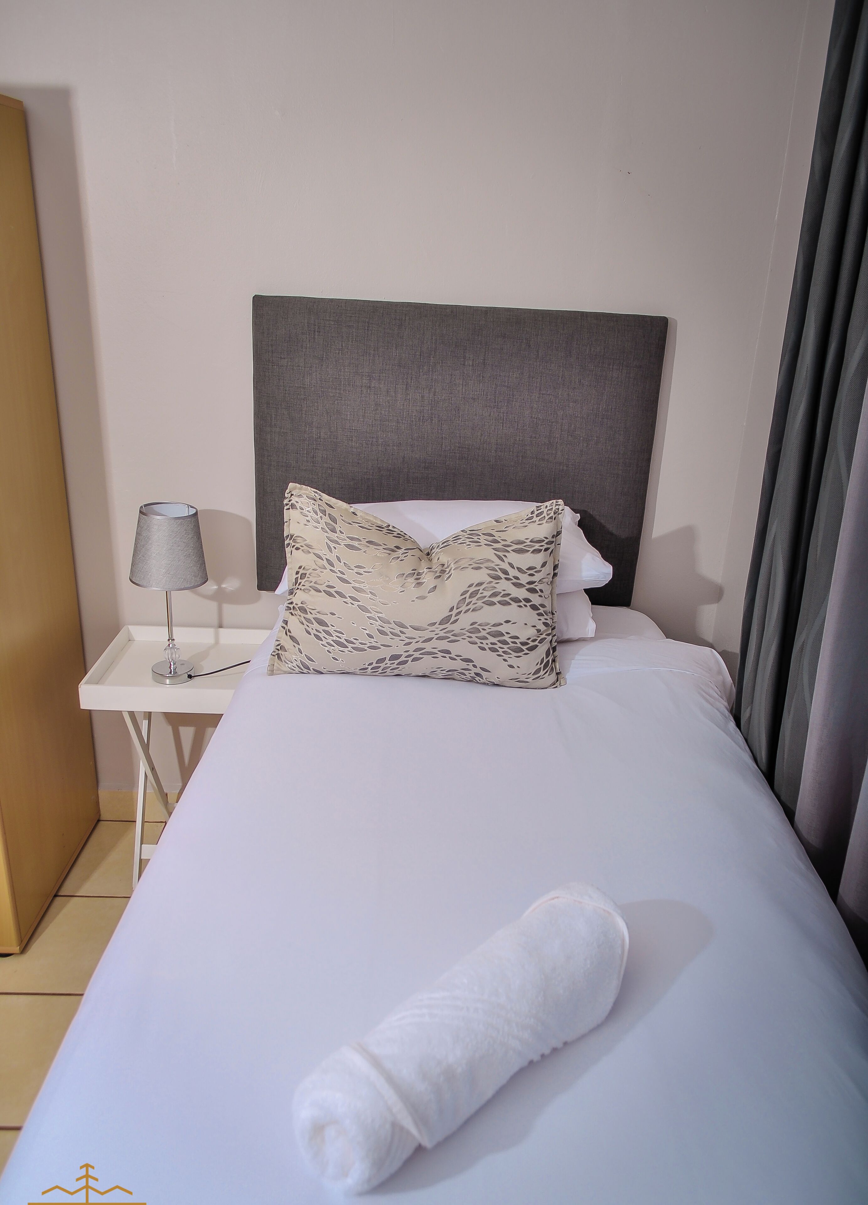 A neatly made bed with a rolled white towel, a patterned pillow, and a plain pillow on a white nightstand with a lamp.