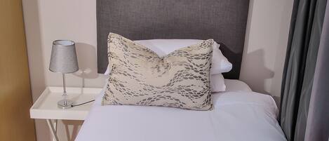 A neatly made bed with a rolled white towel, a patterned pillow, and a plain pillow on a white nightstand with a lamp.