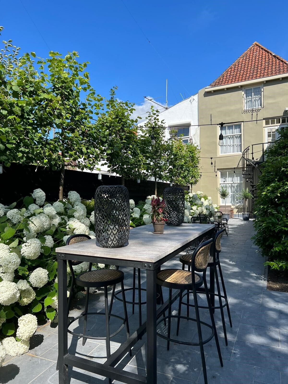 An outdoor patio with a table and chairs, surrounded by white hydrangea bushes and a building with a red-tiled roof.