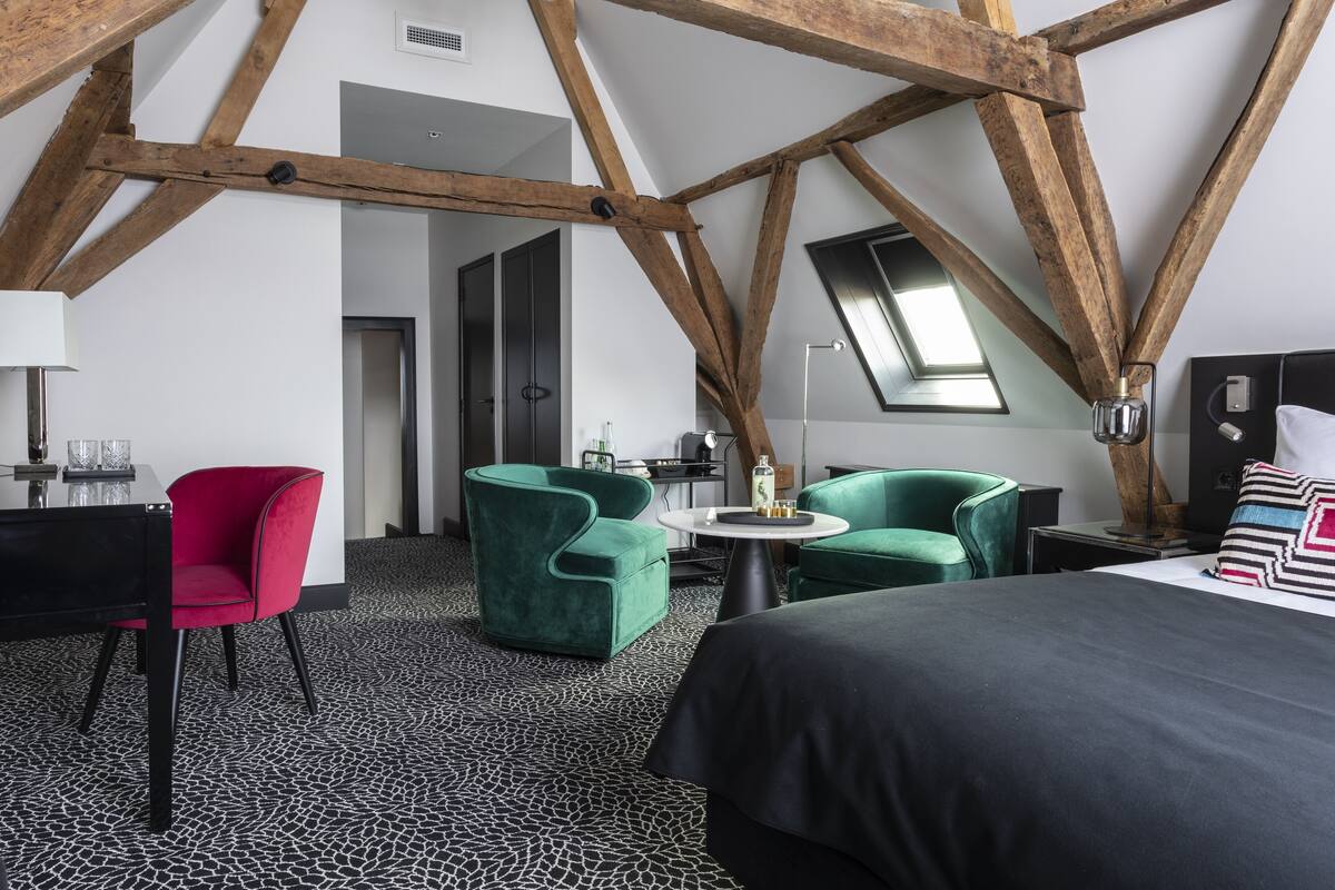 Habitación de hotel moderna con vigas de madera a la vista, suelo con estampado blanco y negro, una cama, dos sillones verdes, una silla roja y una pequeña mesa redonda con una lámpara.