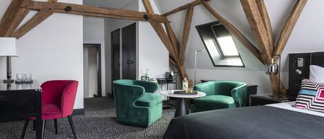 Habitación de hotel moderna con vigas de madera a la vista, suelo con estampado blanco y negro, una cama, dos sillones verdes, una silla roja y una pequeña mesa redonda con una lámpara.