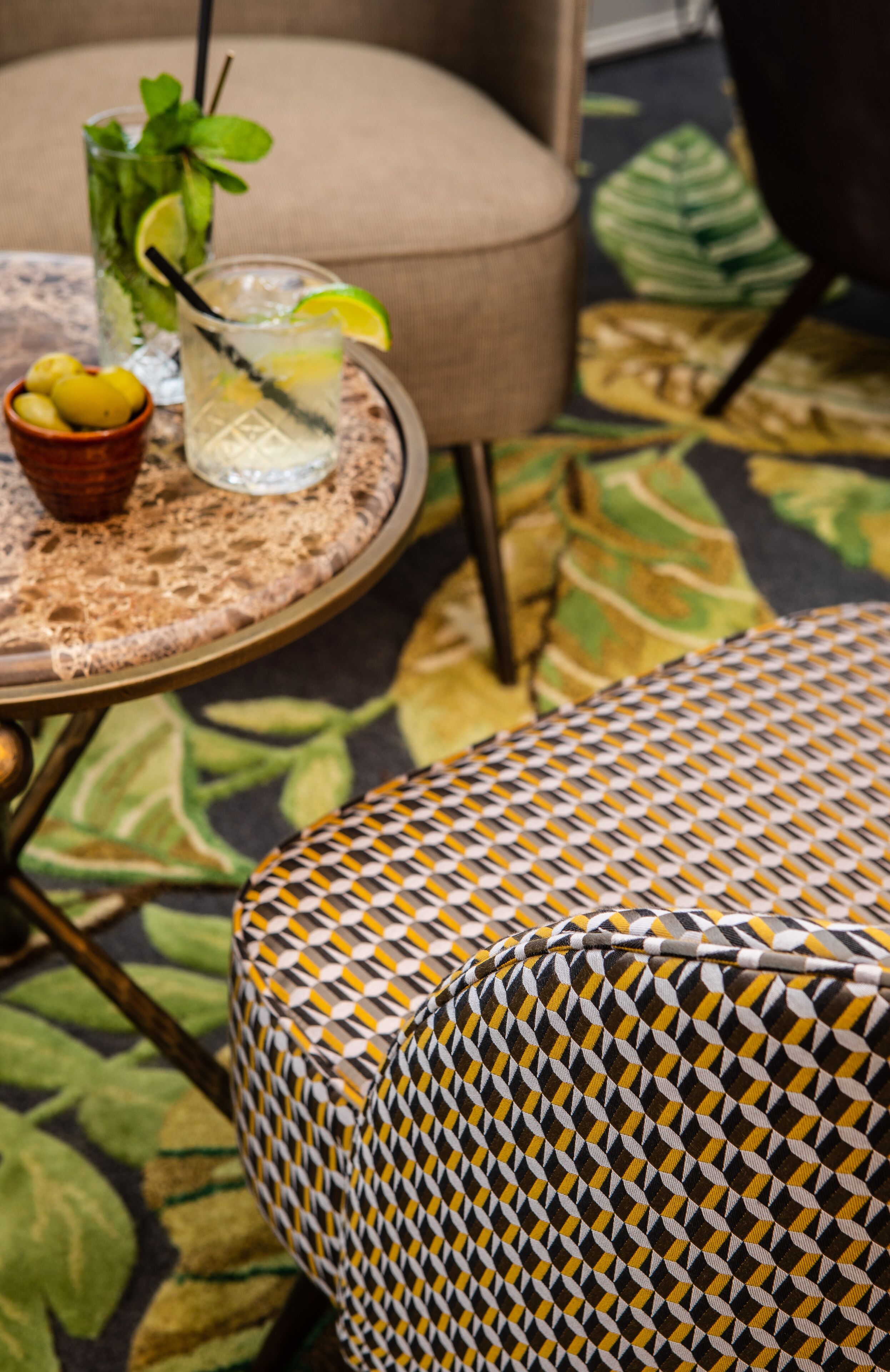 A round table with a glass of iced drink, a bowl of lemons, and a patterned cushion on a floral carpet.