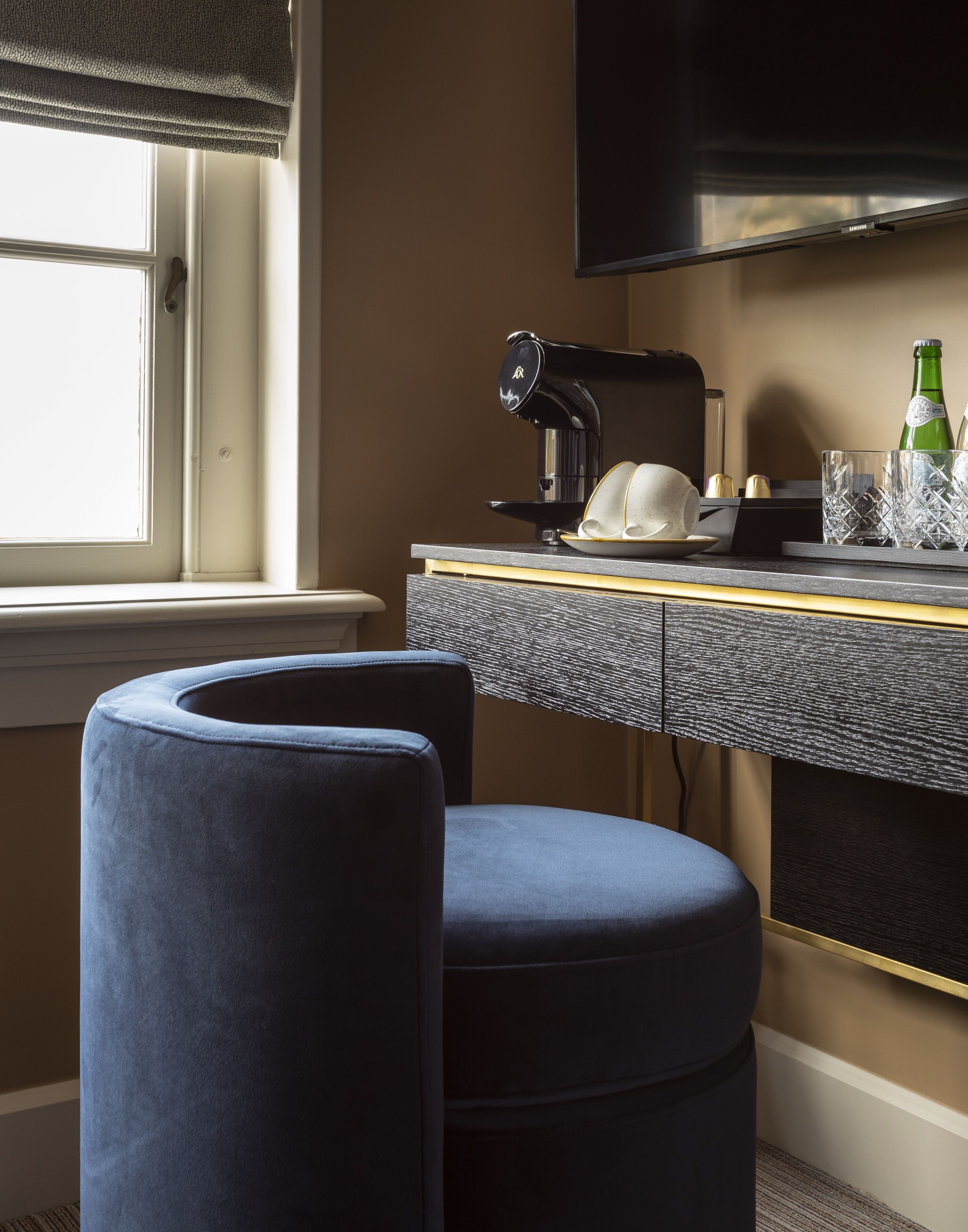 A modern room with a dark grey textured desk, a black coffee maker, a white cup, and a green bottle. There are two blue cushioned stools and a flat-screen TV mounted on the wall.