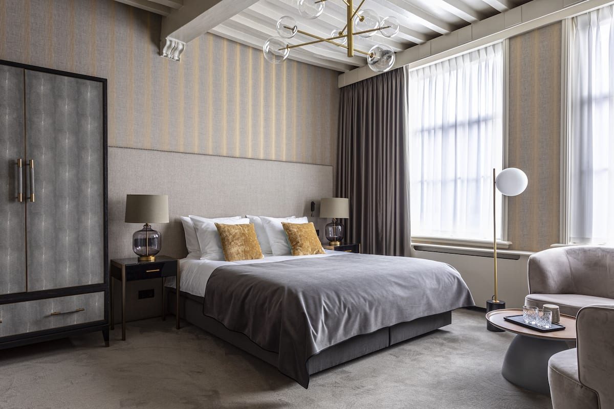 Una habitación de hotel moderna con una cama grande, un armario, una mesita de noche con lámparas y vistas al exterior a través de amplios ventanales.