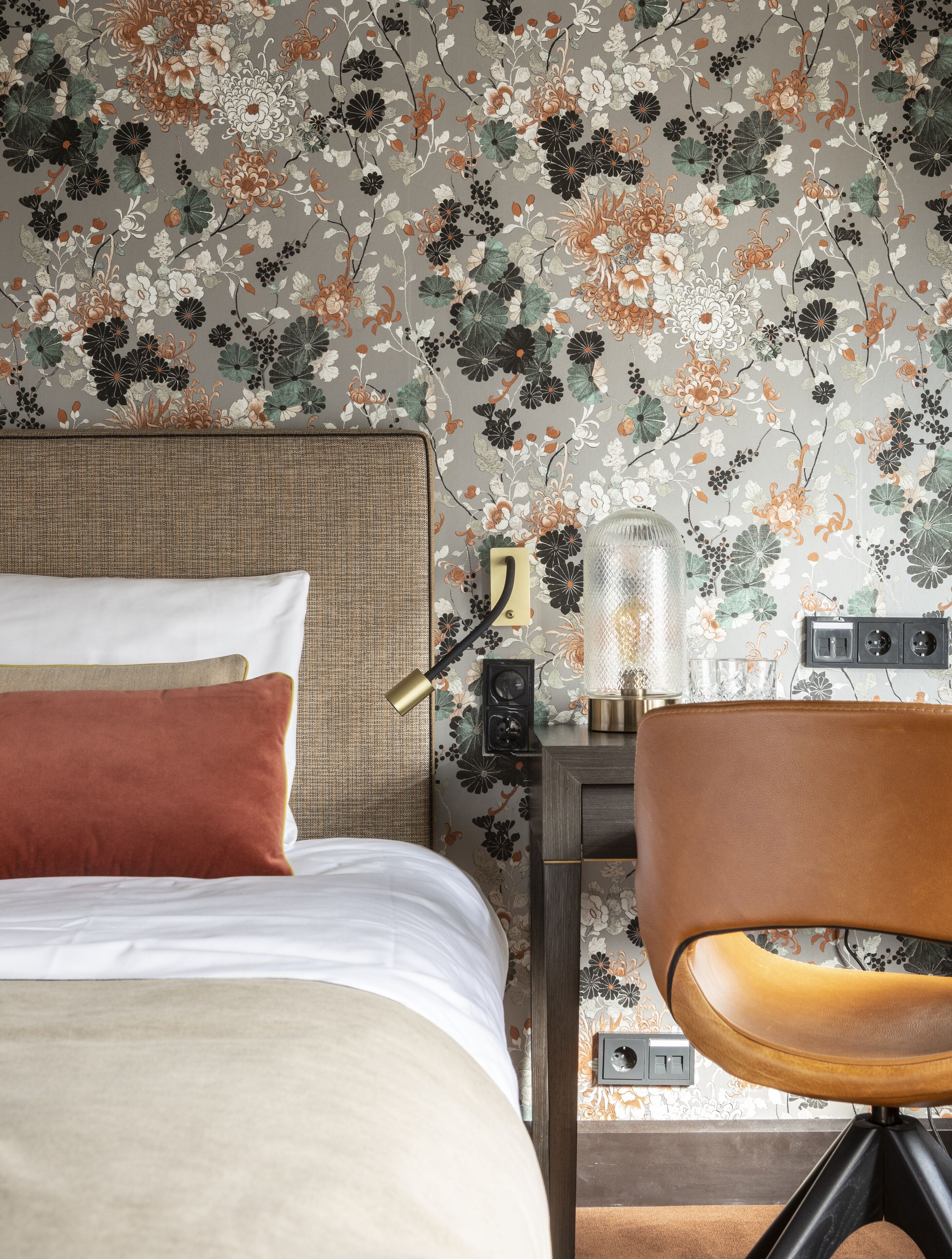 Un dormitorio con papel tapiz floral, una cama con ropa de cama blanca y marrón, una lámpara de noche, una mesita de noche con una lámpara y una silla.