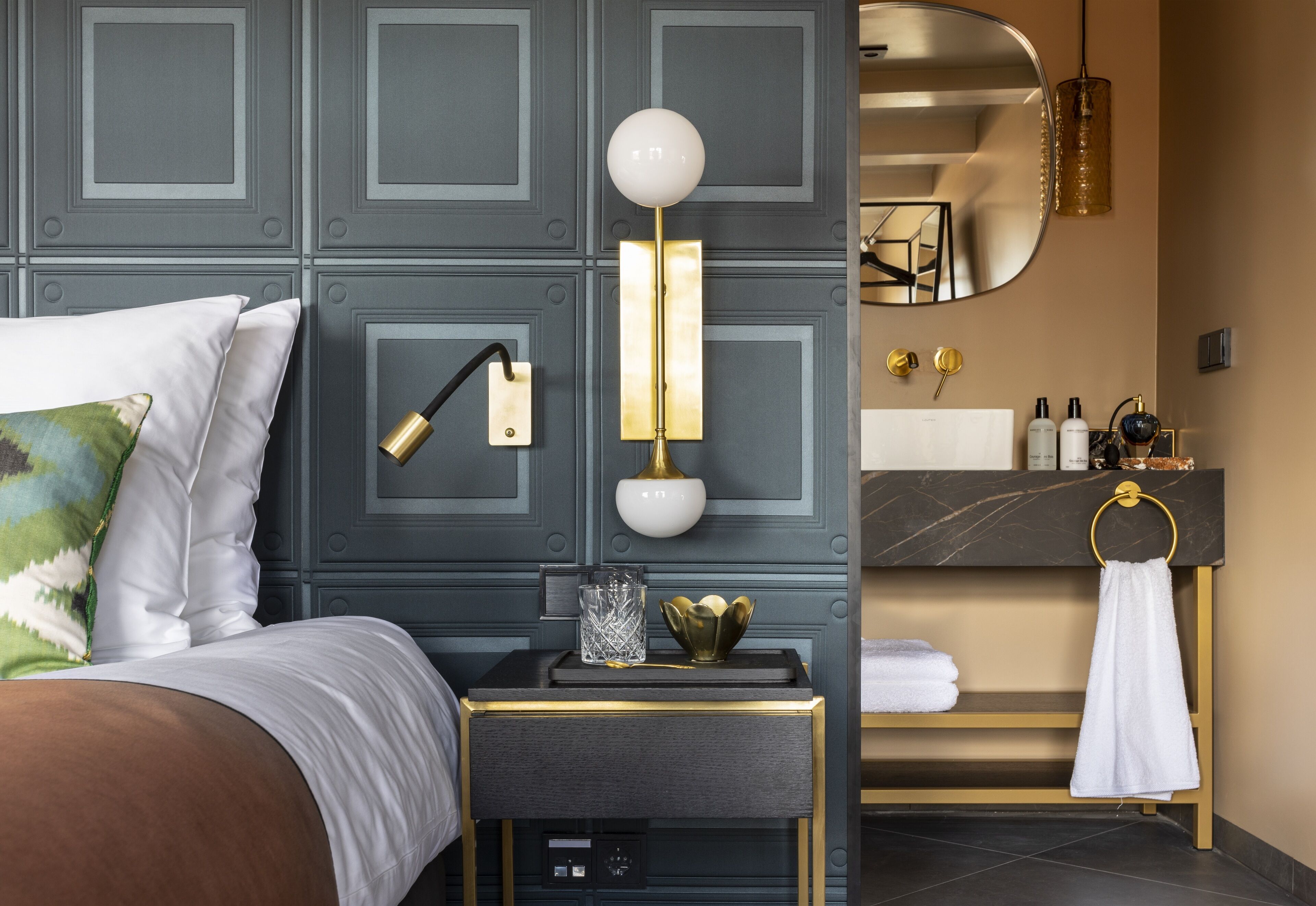 A modern bedroom with a bed, bedside table, lamp, and a bathroom with a sink and towel rack.
