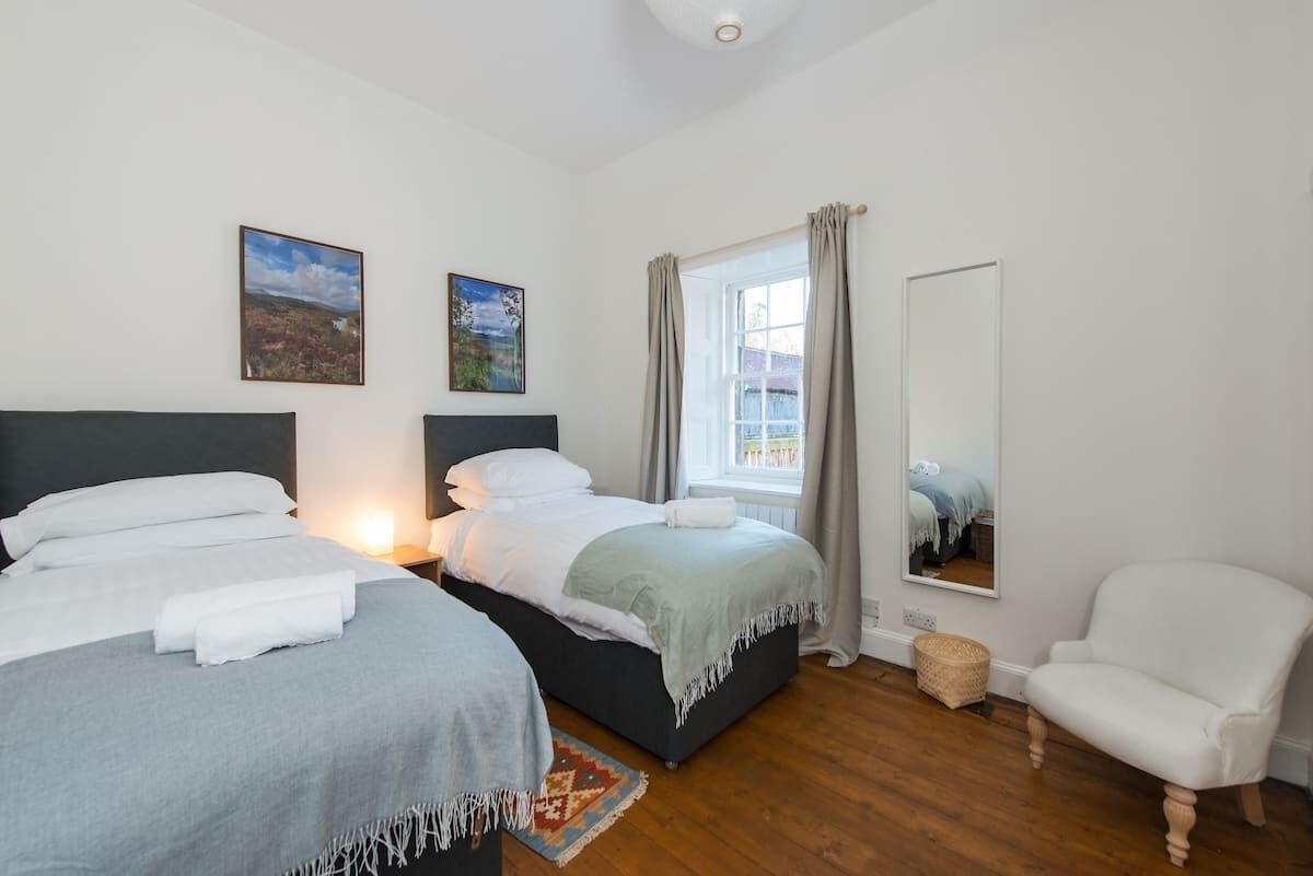 A bedroom with two beds, a window with curtains, a chair, and a mirror.