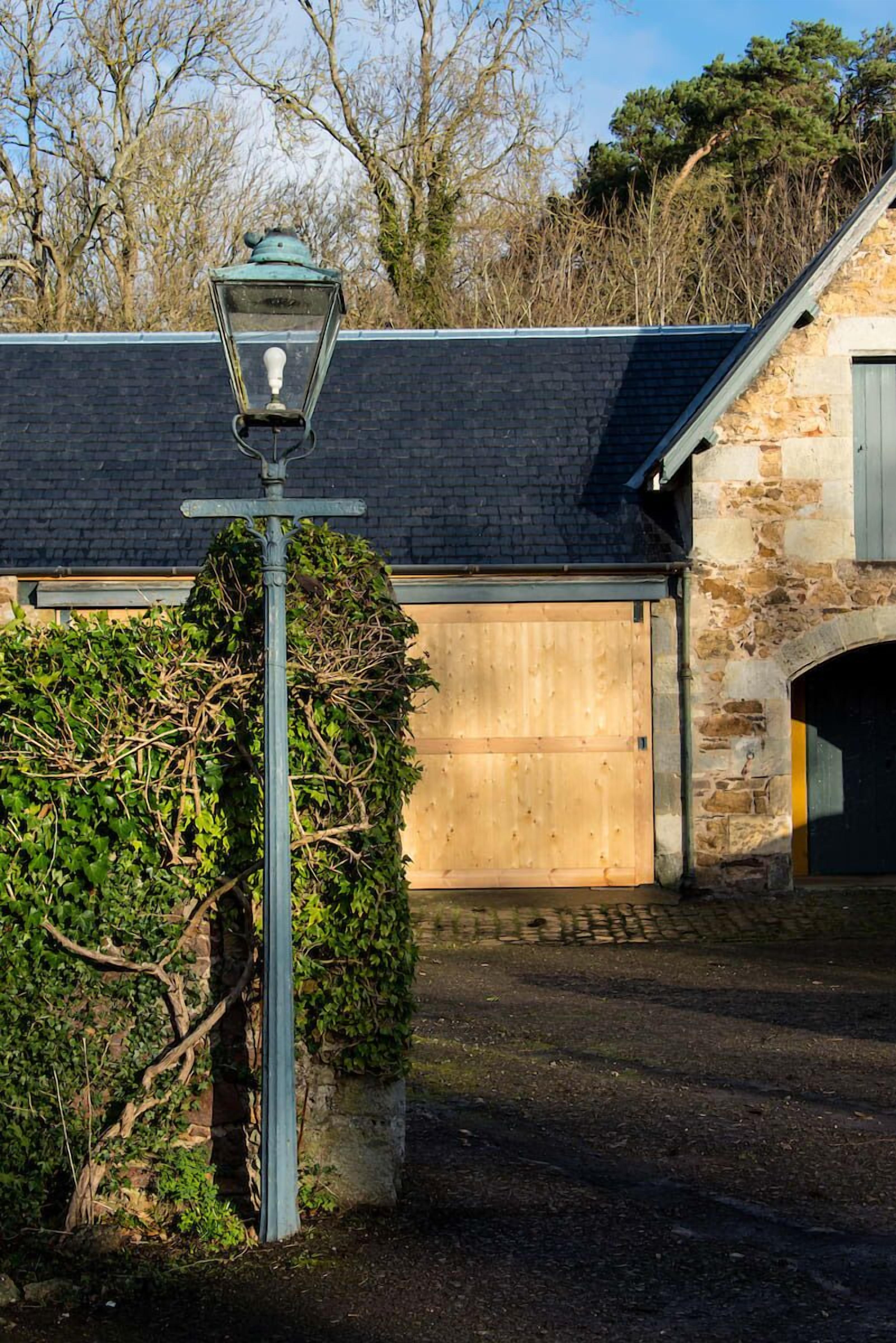 Un bâtiment traditionnel en pierre, avec une porte de garage en bois, un lampadaire en métal et une haie devant.