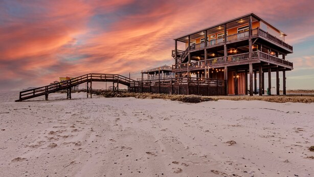 A multi-level wooden structure with a deck and stairs on a sandy beach, under a sunset sky. - Beachfront, Huge Deck and Private Heated Pool (Bolivar Peninsula)