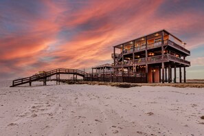 A multi-level wooden structure with a deck and stairs on a sandy beach, under a sunset sky. - Beachfront, Huge Deck and Private Heated Pool (Bolivar Peninsula)