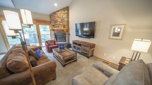A living room with a stone fireplace, a flat-screen TV, and a sofa. - Forested Townhome w/ Garage, Hot Tub & Shuttle (Keystone)