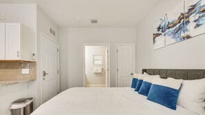 A bedroom with a large bed, white walls, and a gray headboard. There is a painting on the wall and a trash can by the door. - Renovated PCB Studio, 2 Min Walk To Laguna Beach! (Panama City Beach)
