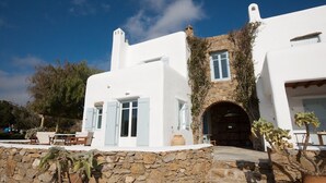 A white building with a stone facade, a wooden door, and a small table with chairs outside. - Architect owned & designed villa (Ano Mera Mykonos)