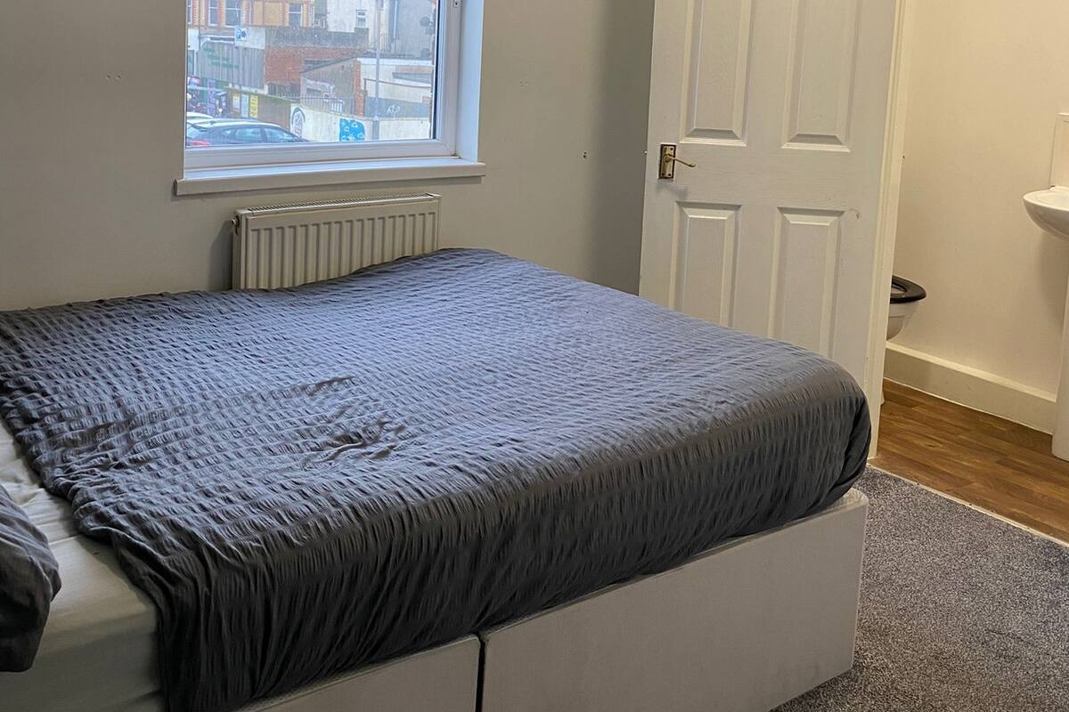 A bedroom with a bed, a window, a radiator, a door, and a bathroom with a sink.