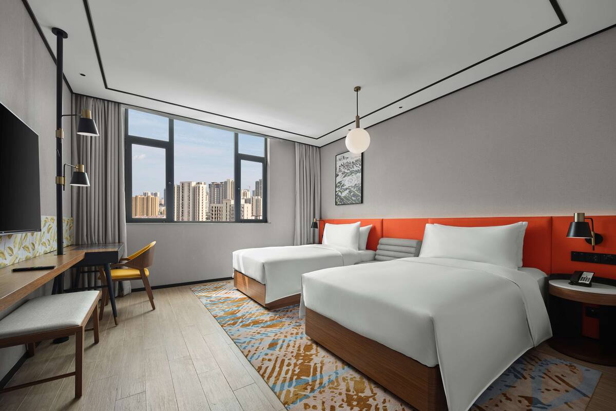 Ein Hotelzimmer mit zwei Betten, einem Schreibtisch, einem Stuhl, einem Fernseher und Blick auf die Stadt.