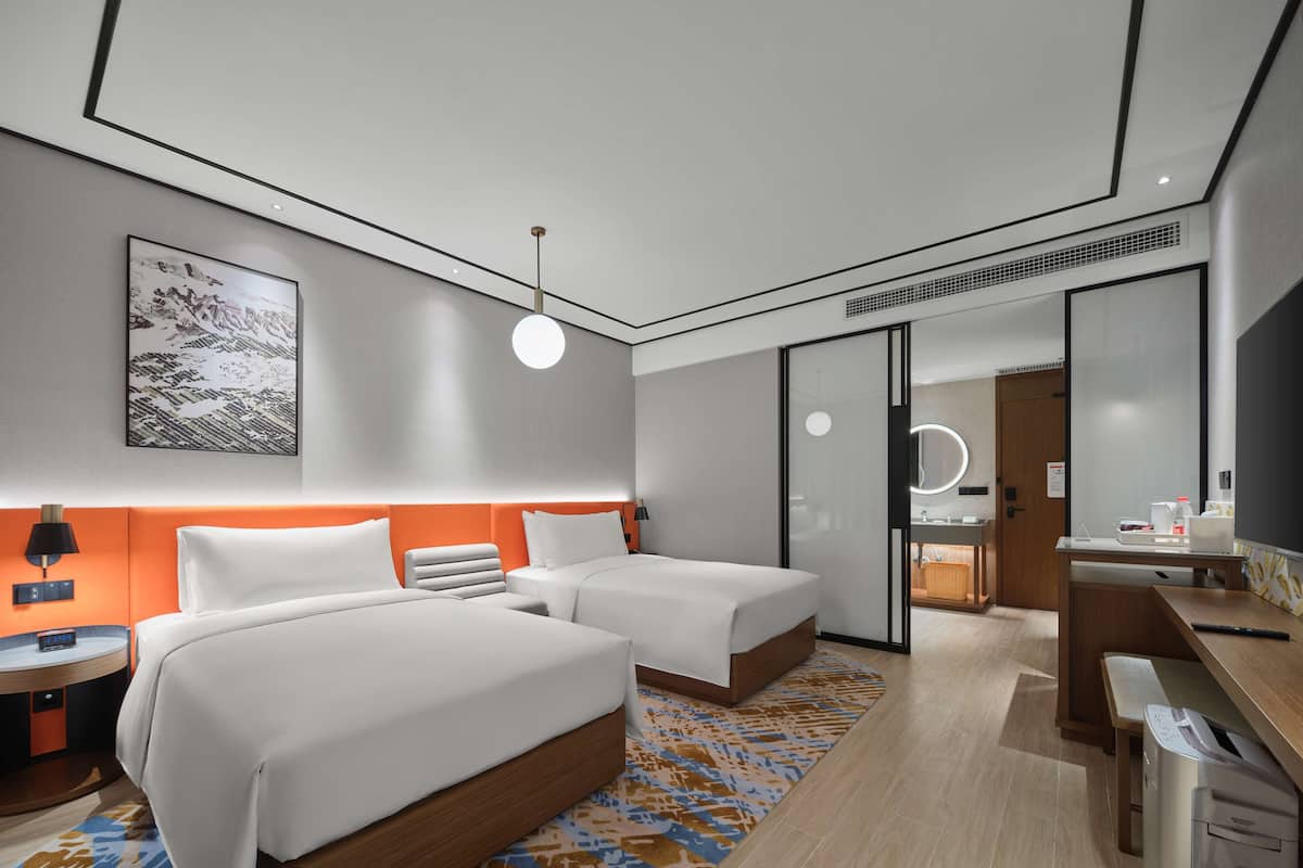 Ein modernes Hotelzimmer mit zwei Betten, einem Schreibtisch, einem Spiegel und einem durch eine Schiebetür einsehbaren Badezimmer.