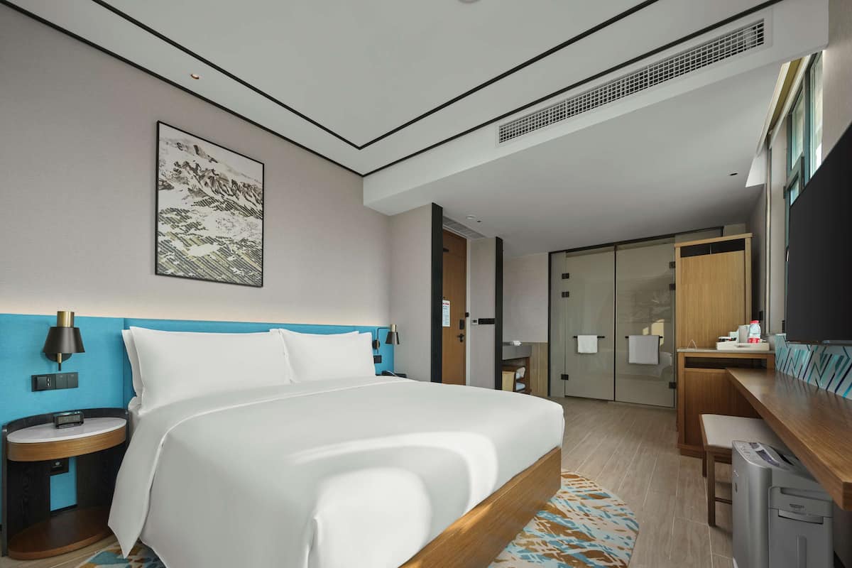 Ein modernes Hotelzimmer mit einem großen Bett, einem Schreibtisch, einem Stuhl, einem kleinen Tisch, einer Lampe, einem Bild an der Wand und einem Schiebetürenschrank.